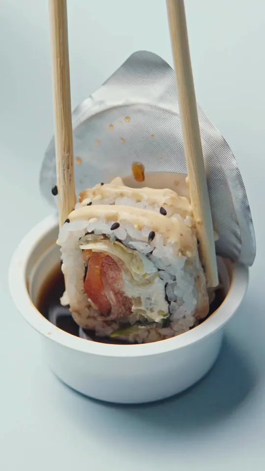 Chopsticks dip a sesame-topped roll into a cup of soy sauce.